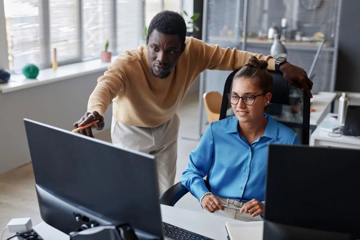 A Man Stands And Points At A Computer Screen Displaying Google Ads, While A Woman In Glasses Sits And Watches Attentively In An Office Setting.