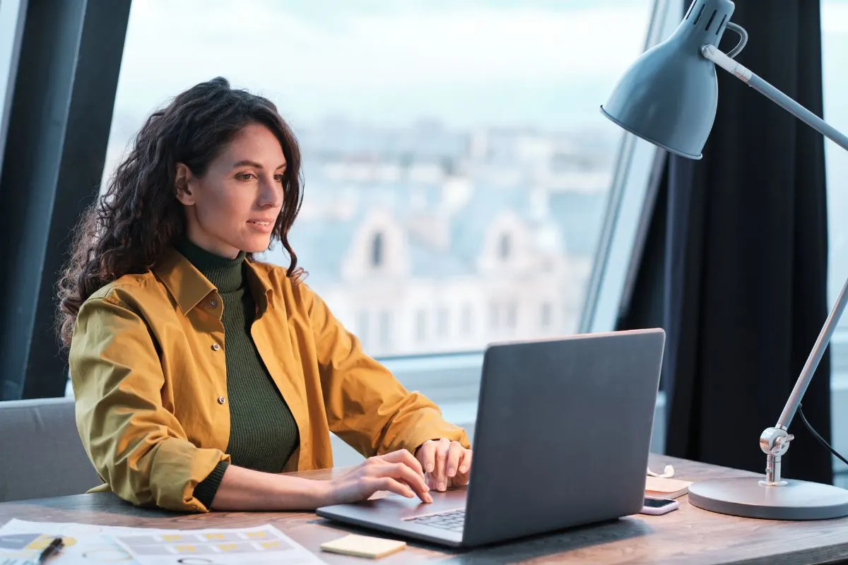 Woman with long curly hair in a yellow jacket works on a laptop at a desk by a window, organizing local citations alongside papers, a pen, and a desk lamp.