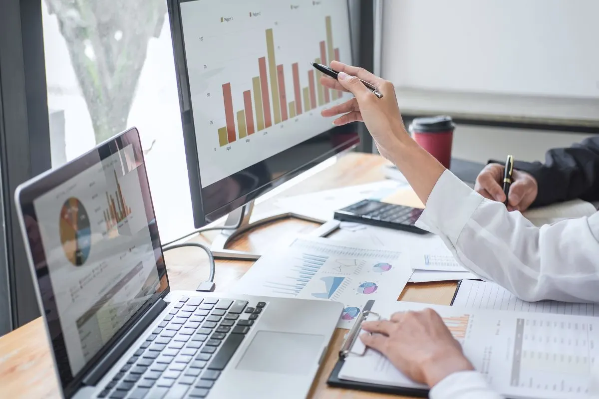 Two people analyze a local SEO report with charts and graphs on computer screens at a desk, alongside a laptop, documents, and a coffee cup.
