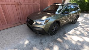 camo green Subaru Outback parked next to a red fence.