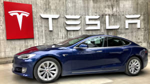 a navy blue Tesla Model S parked in front of a huge Tesla logo hanging on the wall.