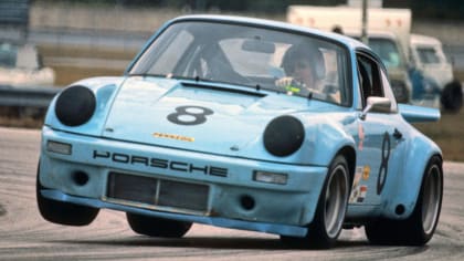 a baby-blue Porsche 911 RSR, with only two wheels on the ground, coming around a corner of a racetrack.