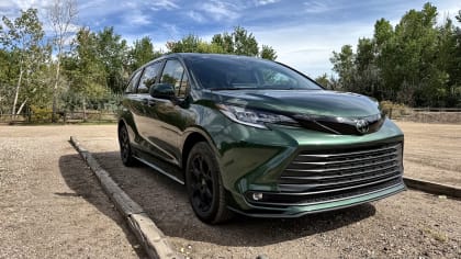 Dark green 2026 Toyota Sienna Woodland Edition photographed from the front three-quarter angle, with black wheels in a wooded parking area.