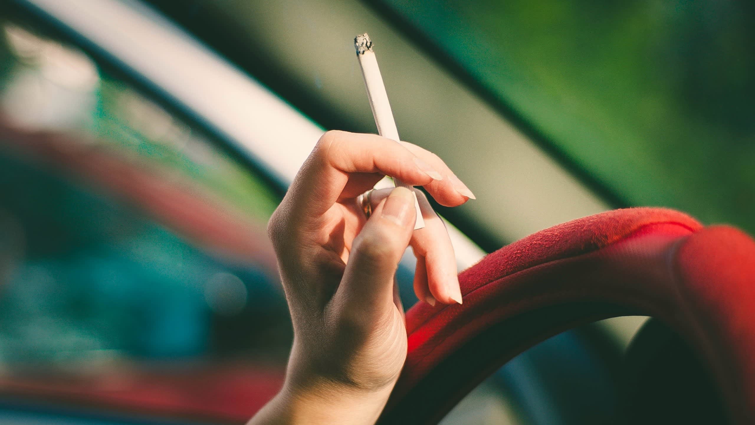 a hand resting on a red steering wheel, holding a cigarette.
