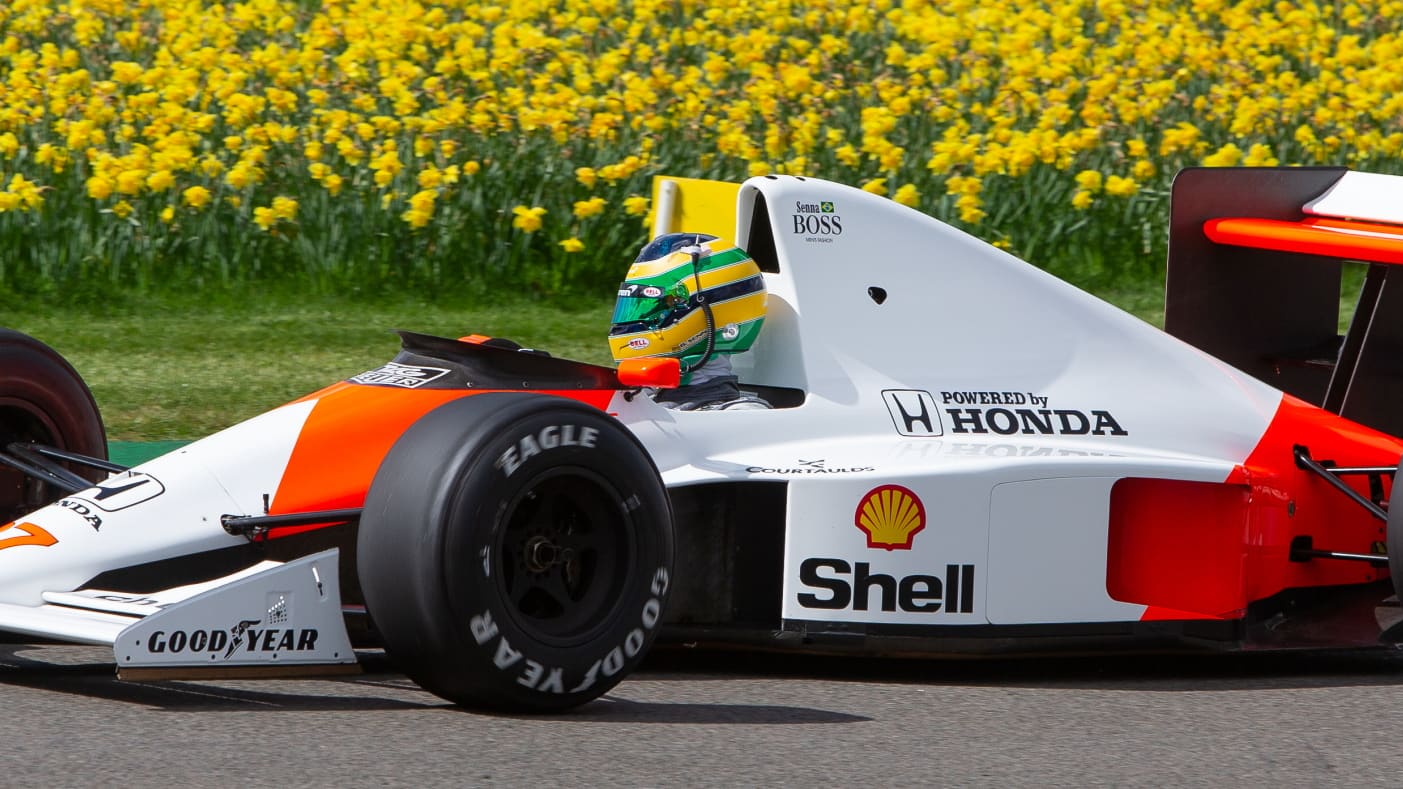 photo of the legendary Ayrton Senna’s 1990 F1 World Championship Mclaren racecar on a race track with a field of yellow flowers.