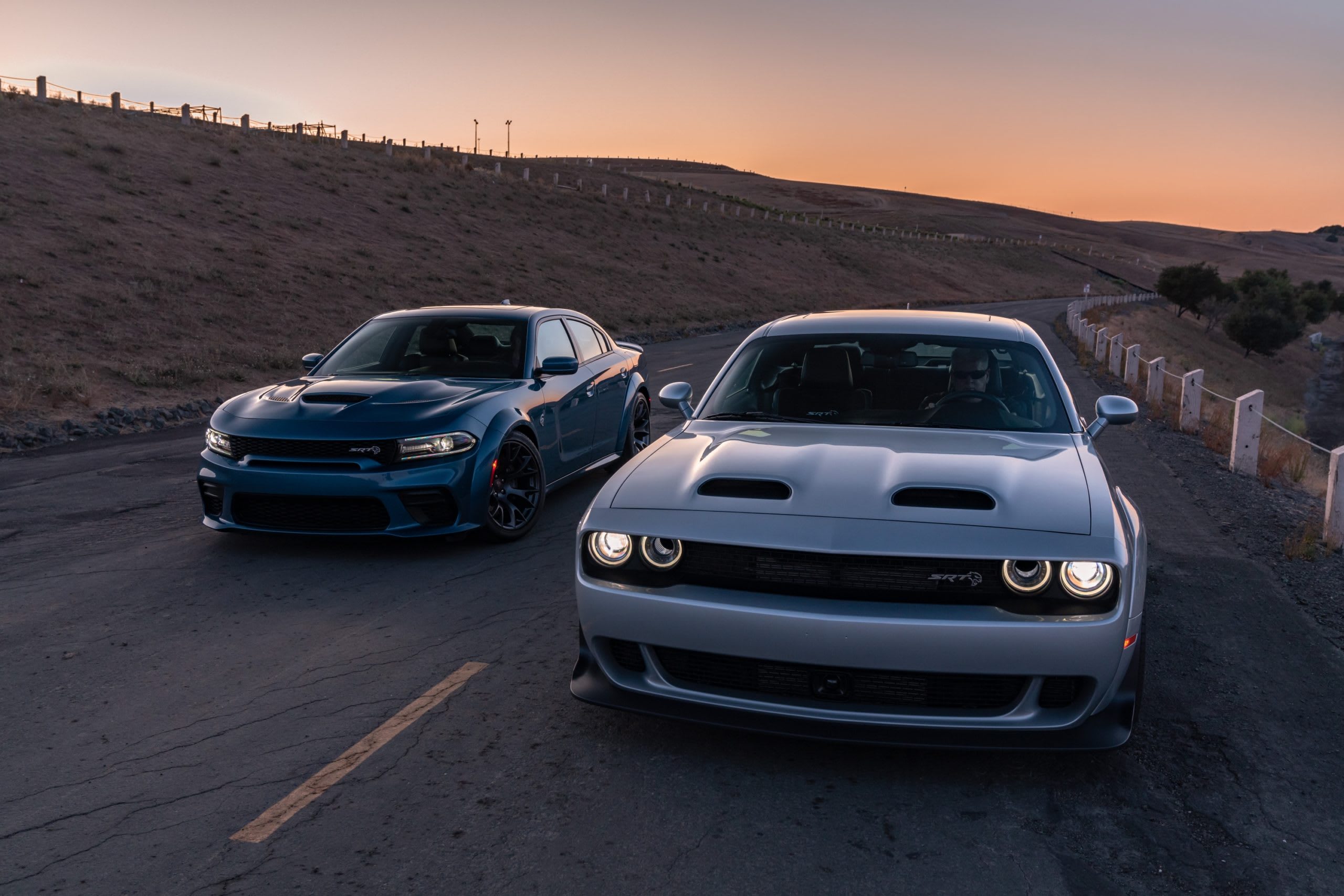 blue and silver Dodge vehicles driving down a road atop a cliff with the ocean in the background.