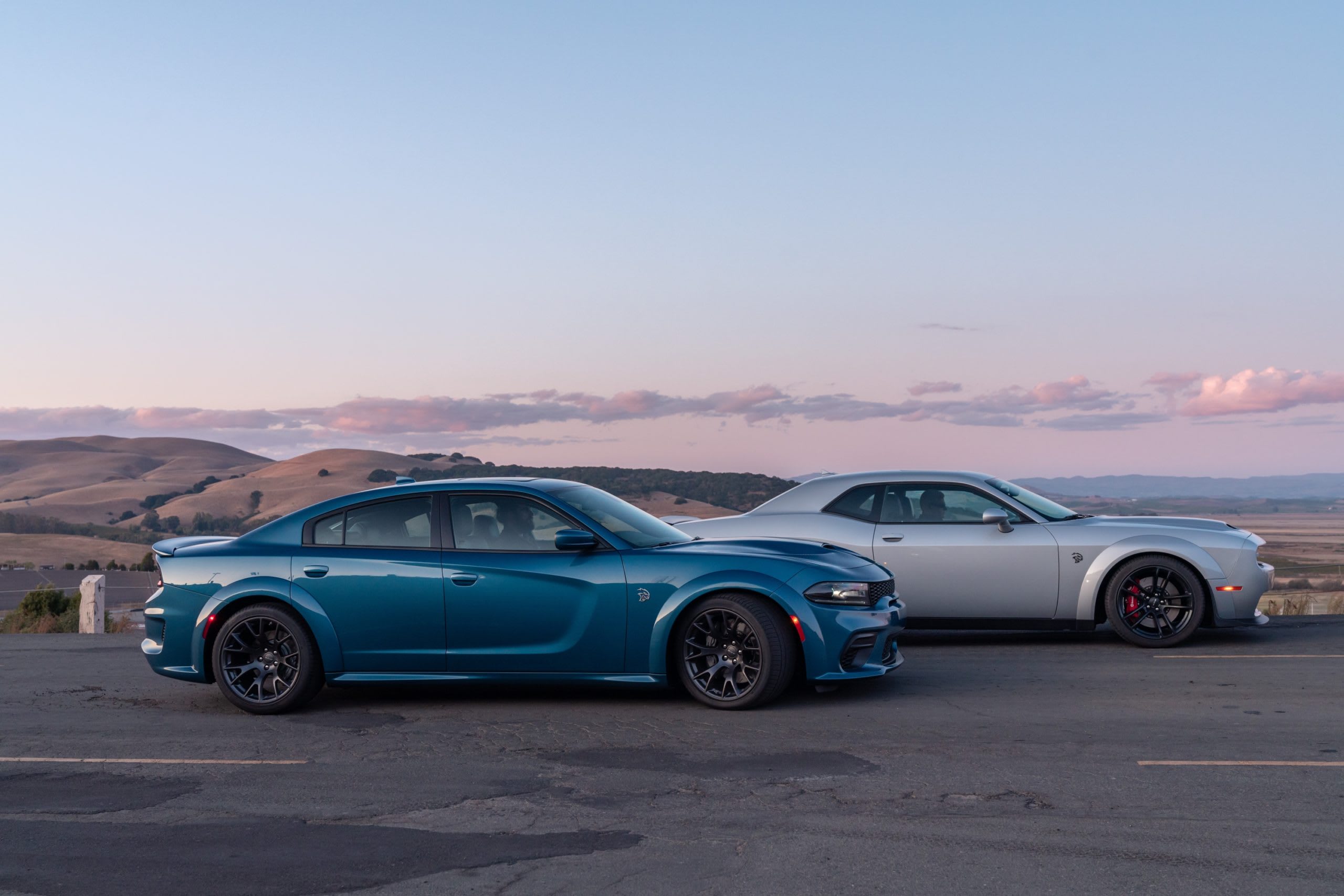 a blue 2020 Dodge Charger SRT Hellcat Widebody and a silver 2020 Dodge Challenger SRT Hellcat Widebody parked next to each other atop a cliff with the ocean in the background.