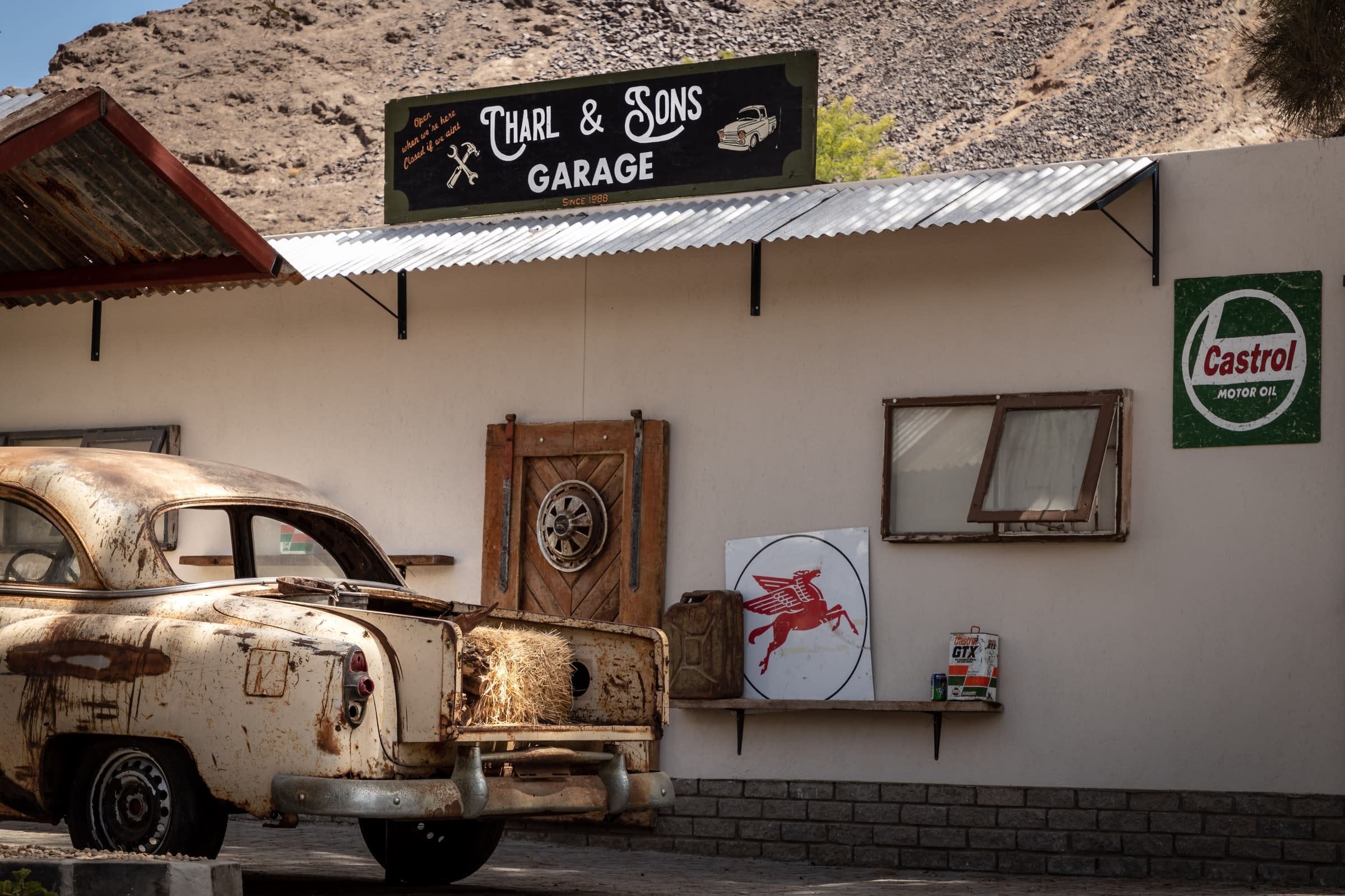A custom garage sign hanging on a rustic house with a rusted out car beside.