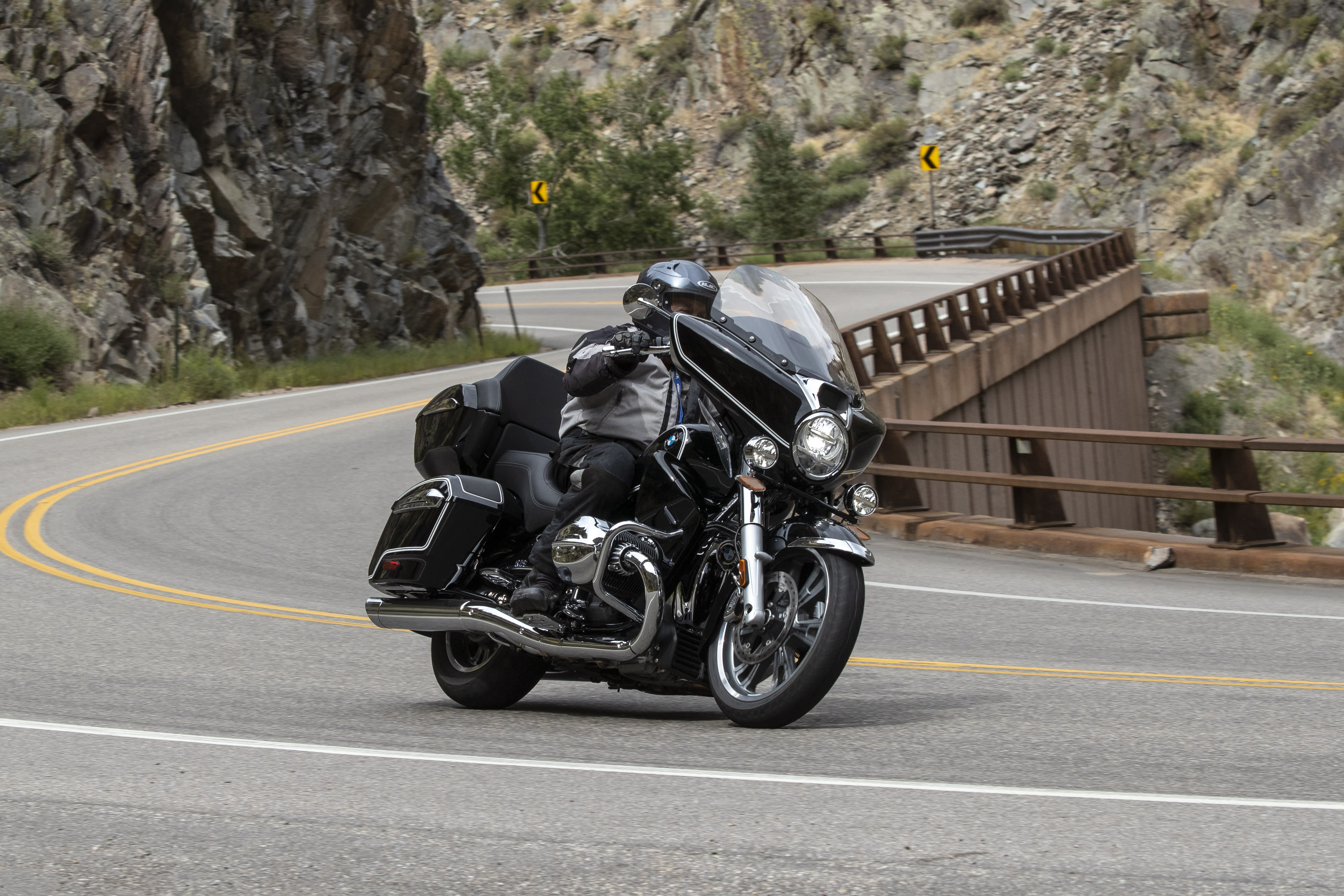 A BMW R 18 Transcontinental cornering on a mountain-side highway with cliffs on one side and a drop on the other.