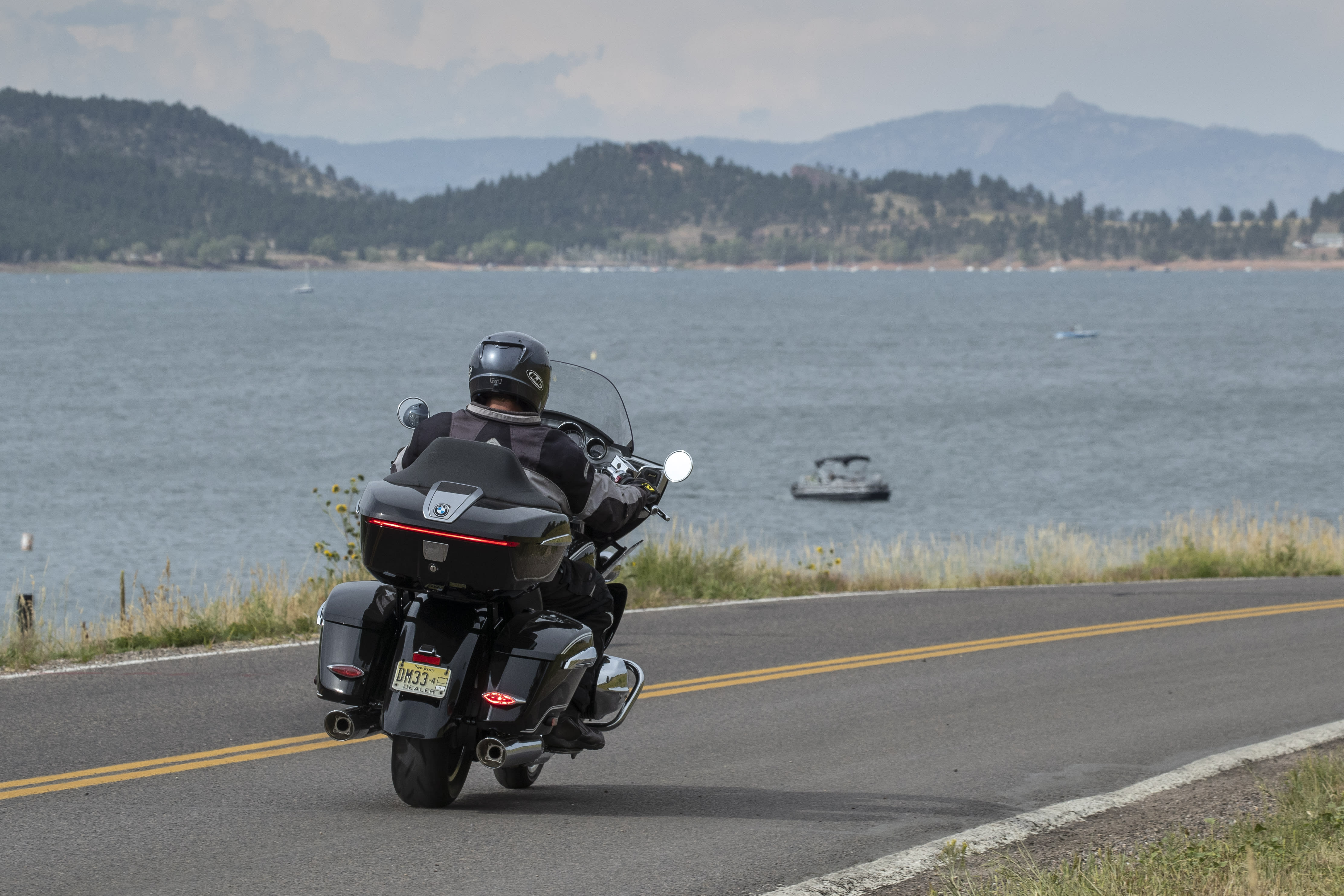 A BMW R 18 driving away from the camera along a lake-side road, with mountains in the background.