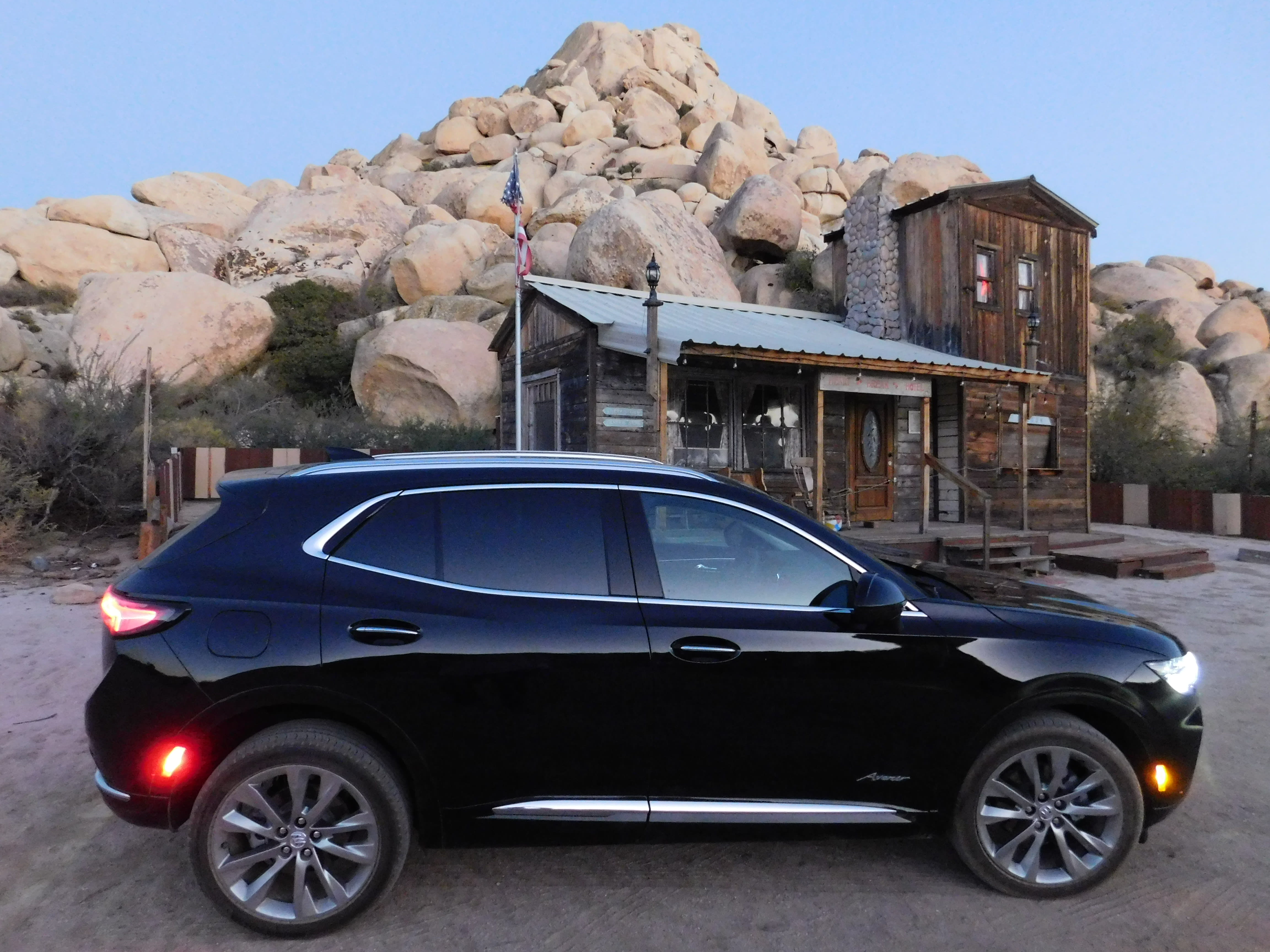 A dark 2022 Buick Envision Avenir parked on desert dirt beside a rustic tavern and with a pile of desert stones piled high in the background.