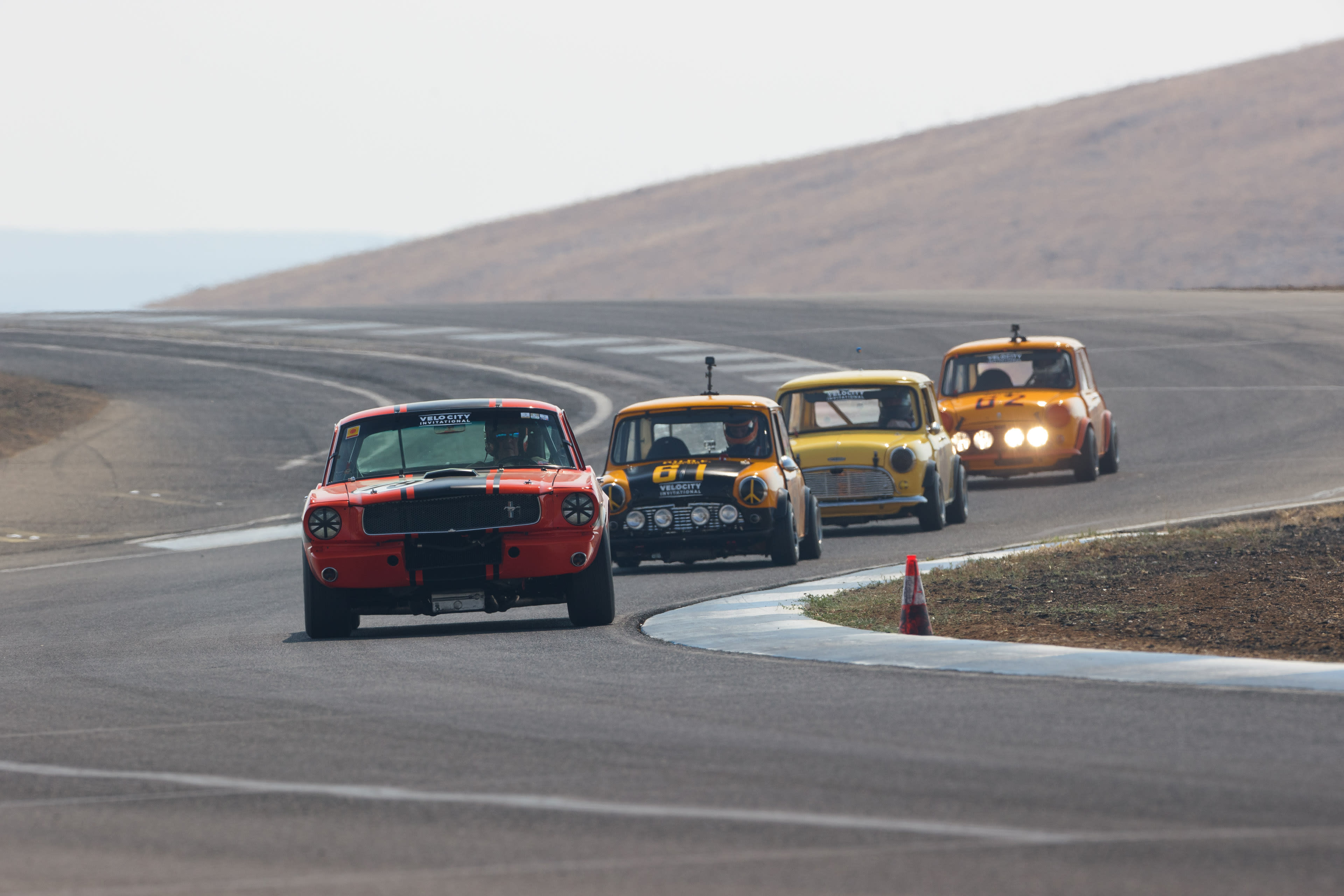 a few vintage (and not) race cars coming around corners at a race track in the desert - with a mustang in the foreground.