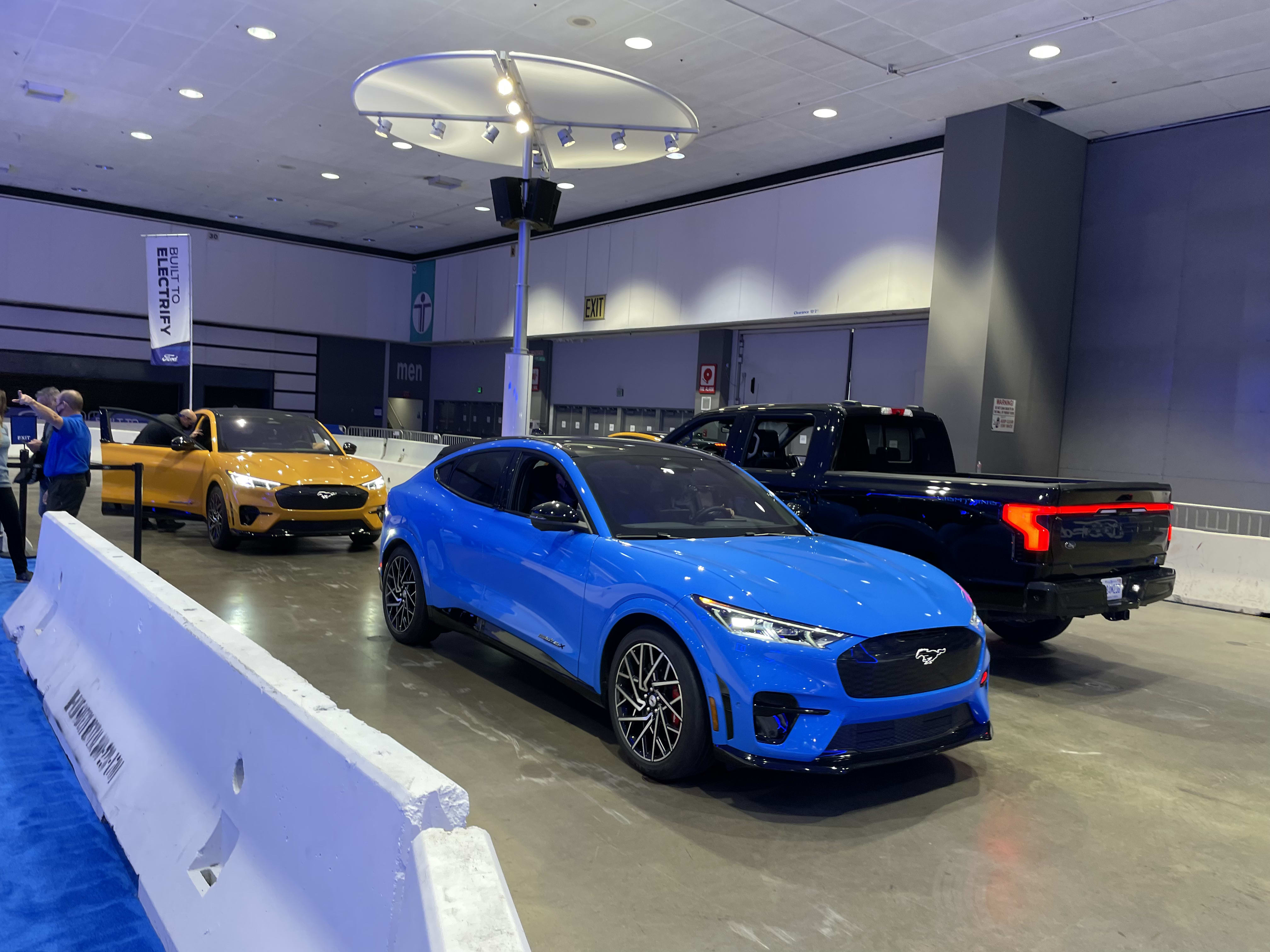 a blue Ford Mustang Mach E heading out on the mini-track at the Los Angeles Auto Show - with a yellow Mach E behind it in line and a pickup truck heading the other way.