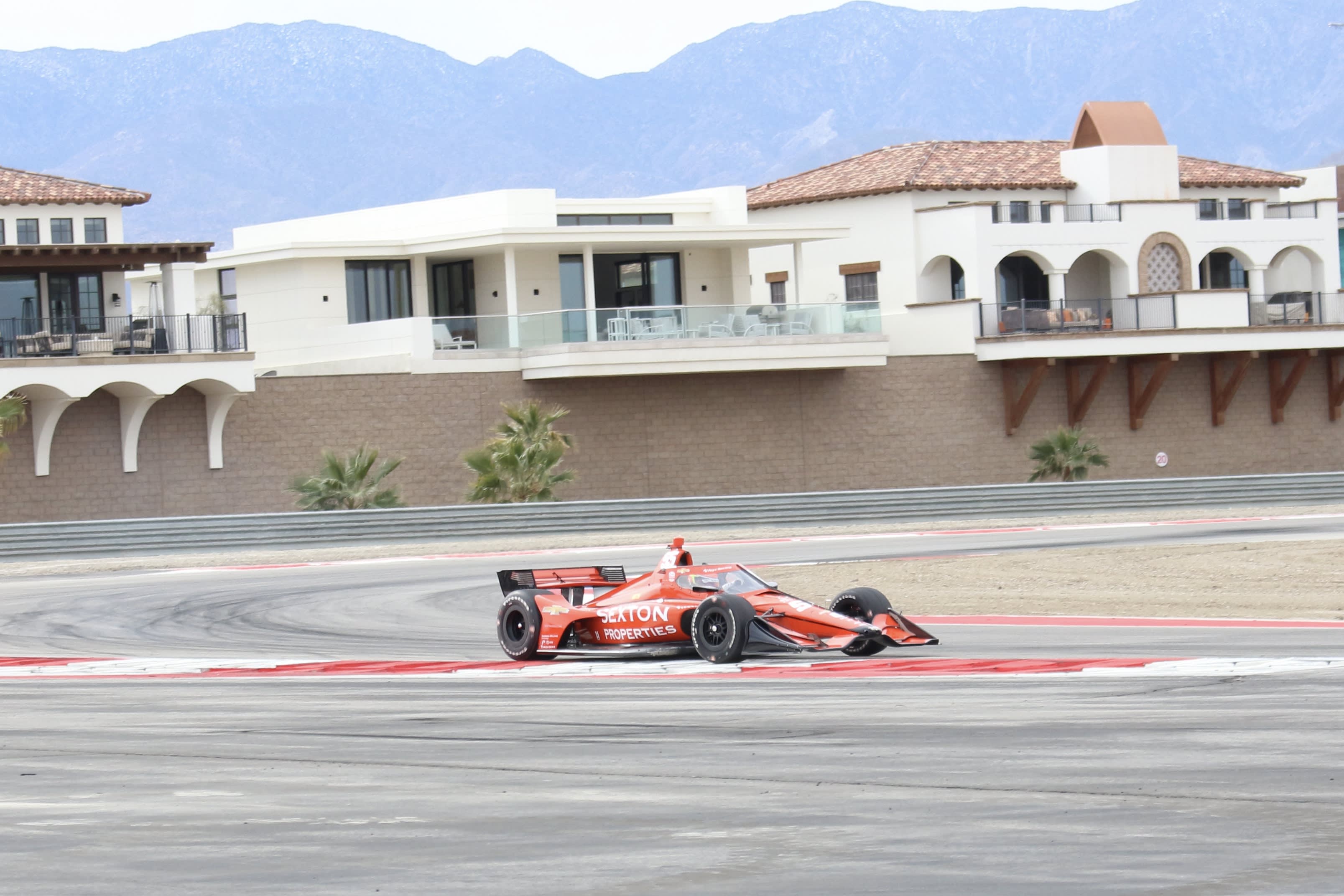 An IndyCar race car coming around a turn on the track at The Thermal Club with the private villas seen in the background.