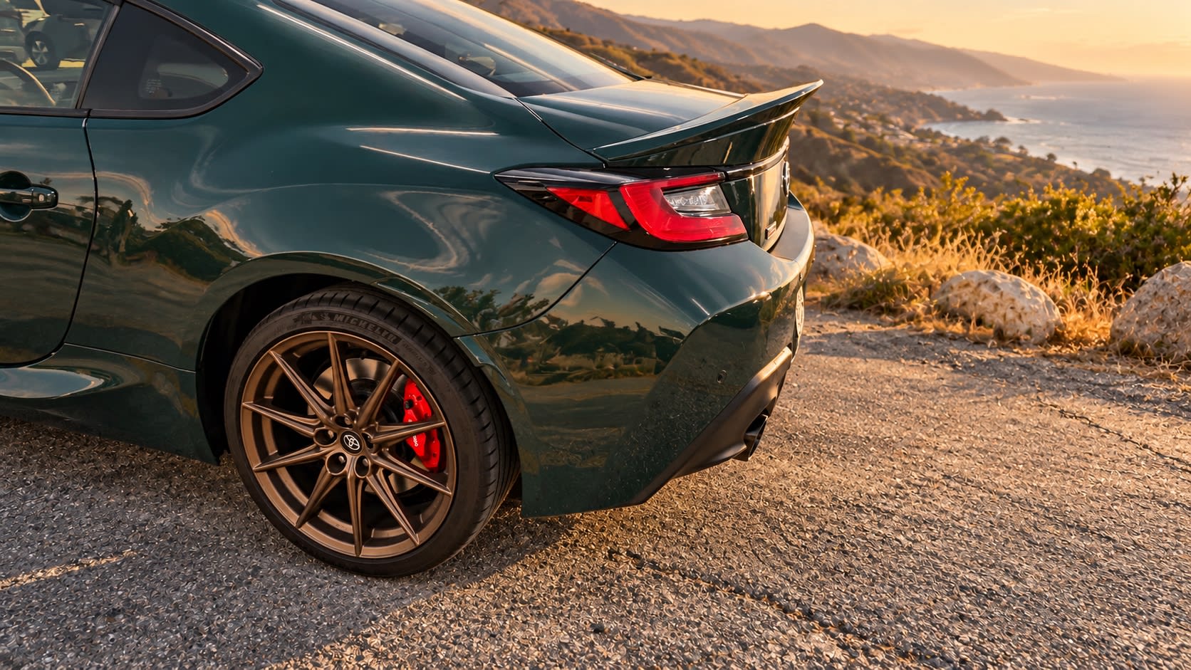 Digitally altered 2025 Toyota GR86 rear-quarter detail showing a bronze wheel, red brake caliper, taillight, and spoiler with a generated coastal background.