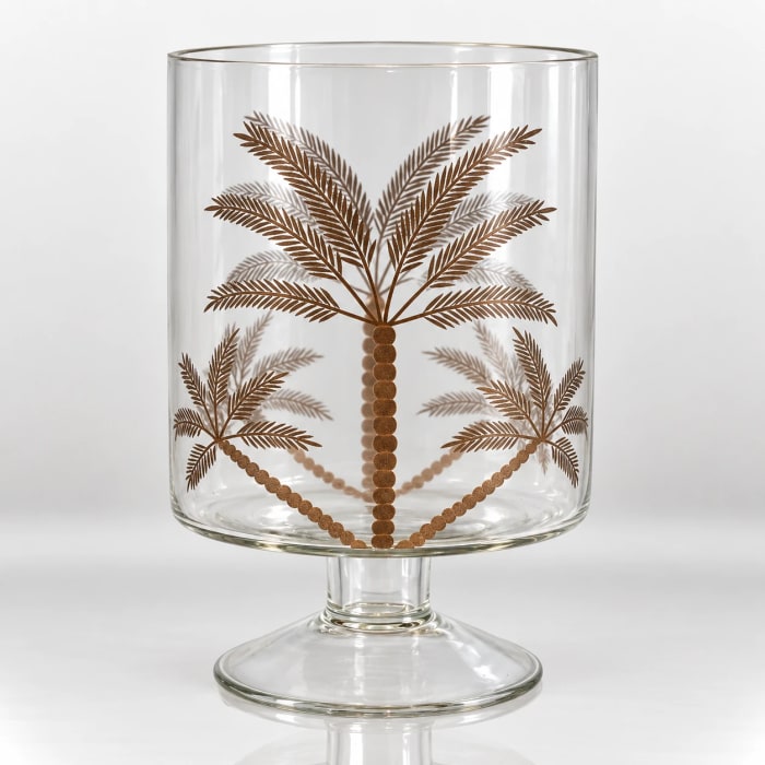 Clear glass pedestal candle holder decorated with a bronze-gold palm leaf botanical design.