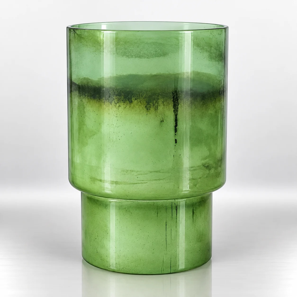 Green glass candle holder with a tall cylindrical upper section, raised base and distressed finish.