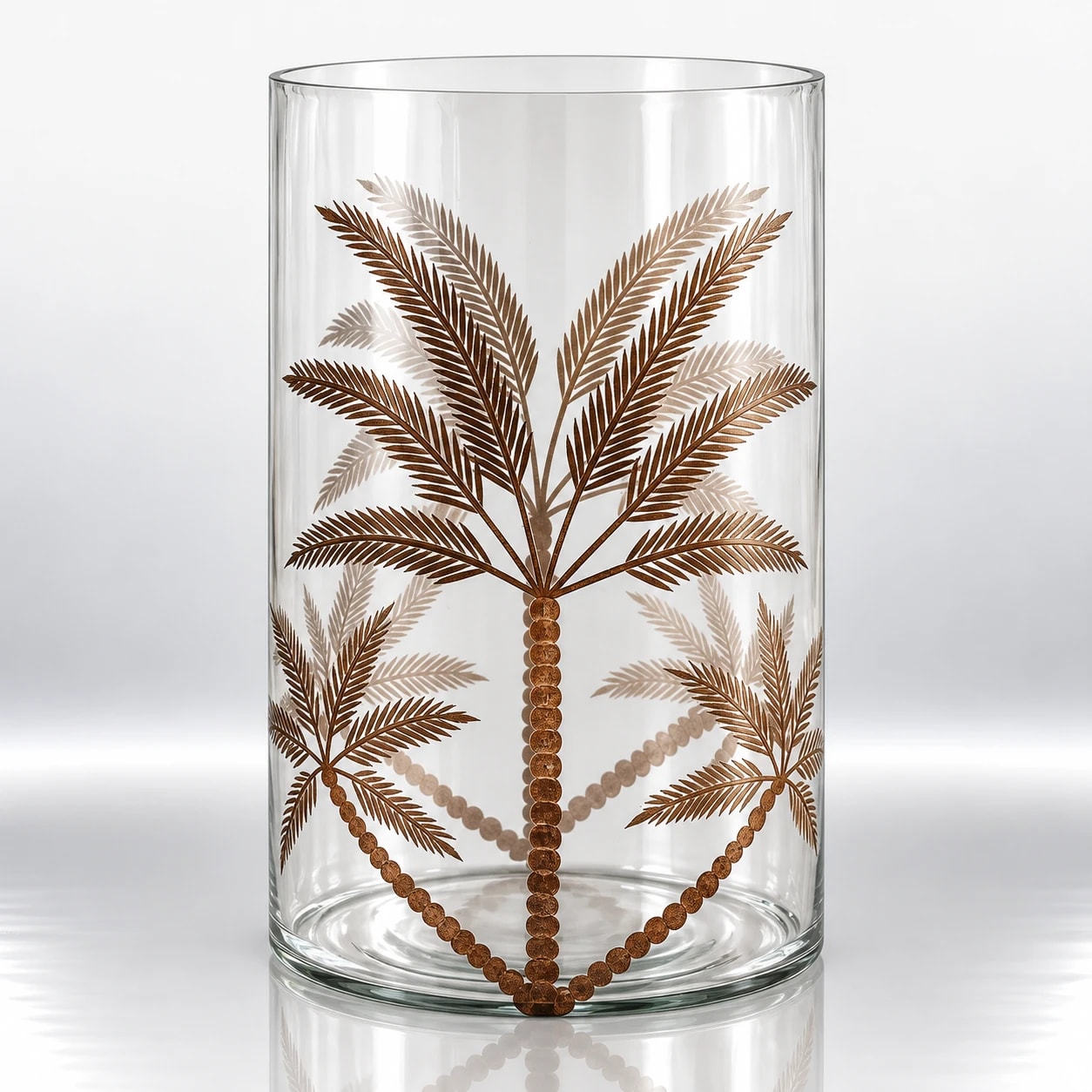 Clear cylindrical glass candle holder decorated with bronze palm trees, detailed foliage and dotted trunk patterns.