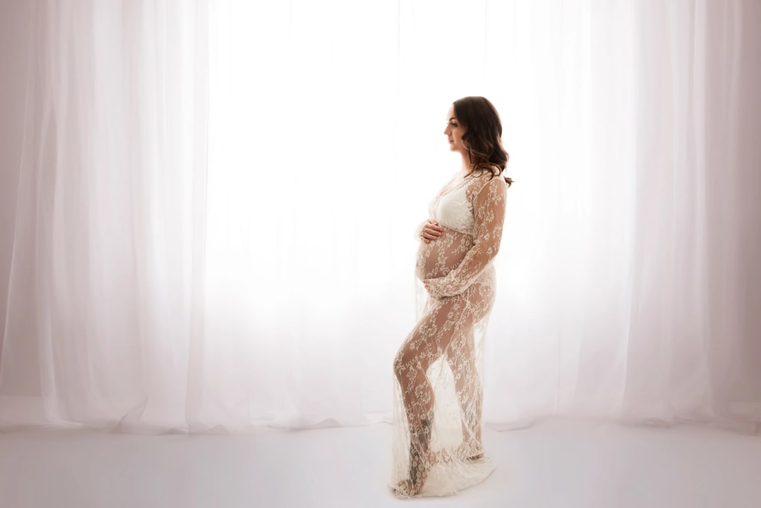 Pregnant woman in lace gown stands by a window, embracing her belly. Maternity photography session in East Kilbride