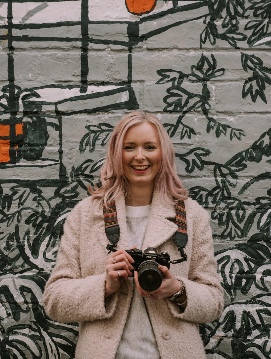 Woman holding a camera in front of a mural backdrop, smiling warmly