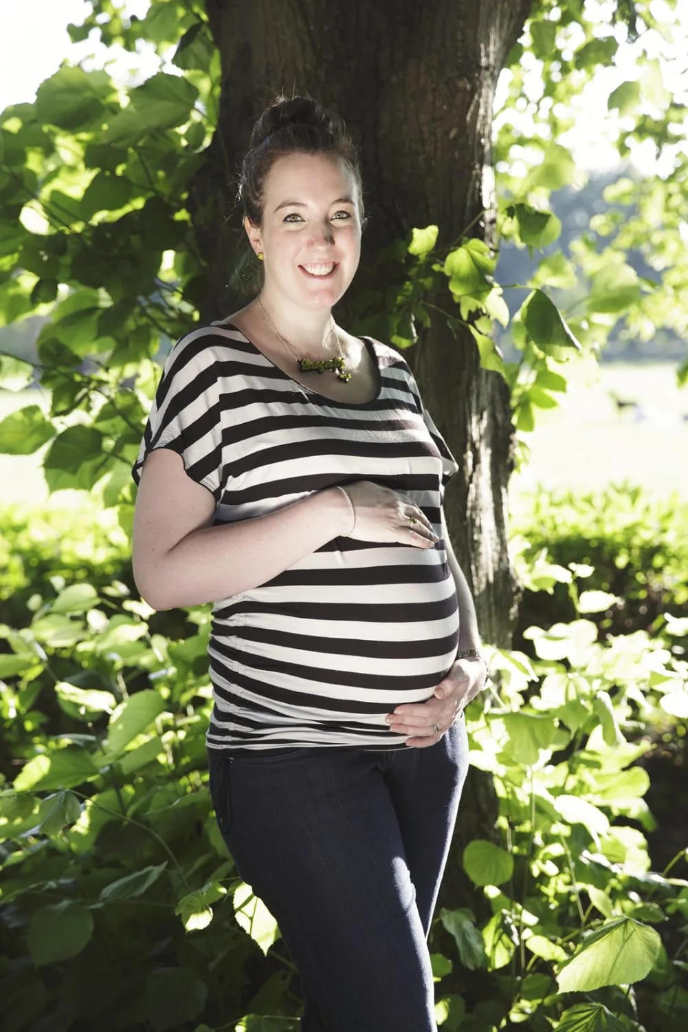 A pregnant woman in a striped shirt holding her belly, standing under a tree during a Maxwell Park maternity session in Glasgow.