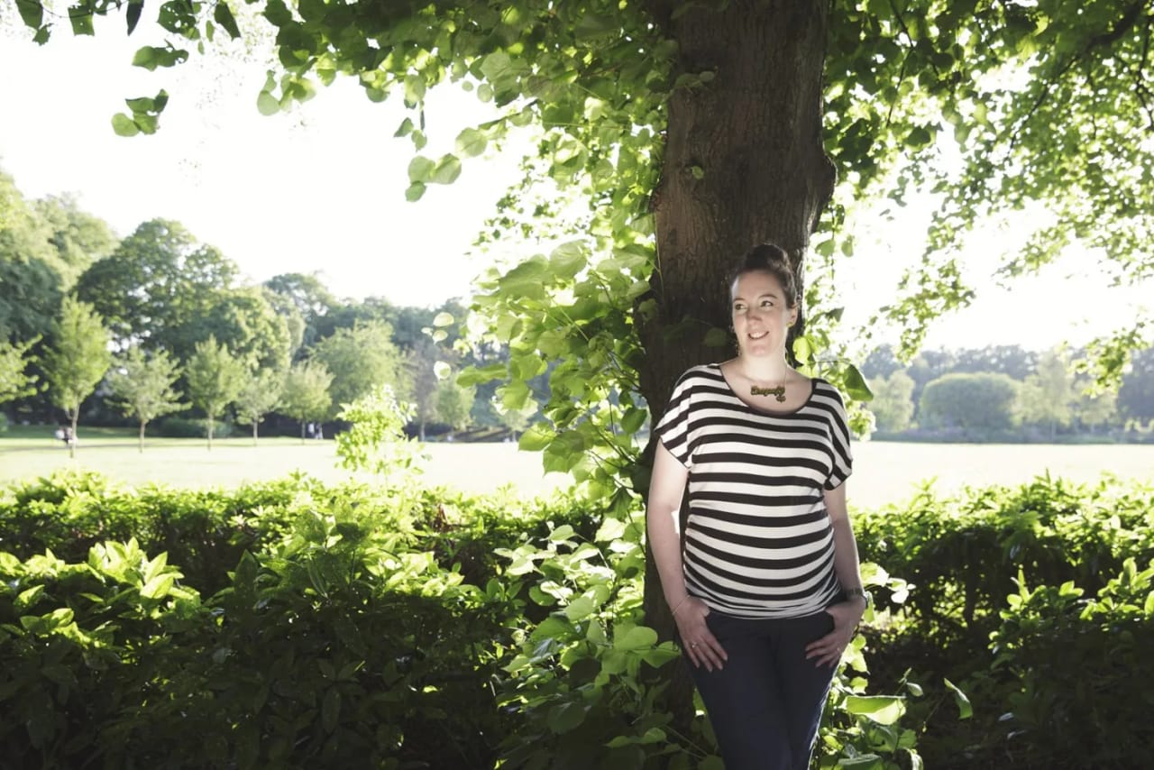 A maternity session in Maxwell Park, Hamilton, featuring a pregnant woman in a striped shirt standing by a tree in the sunshine.