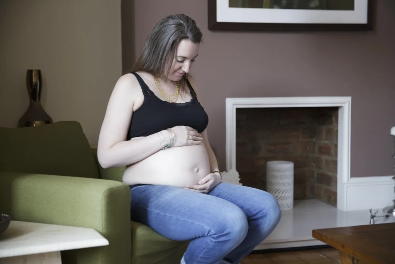 A pregnant woman sitting on a green chair, gently cradling her belly, during a Maxwell Park maternity session