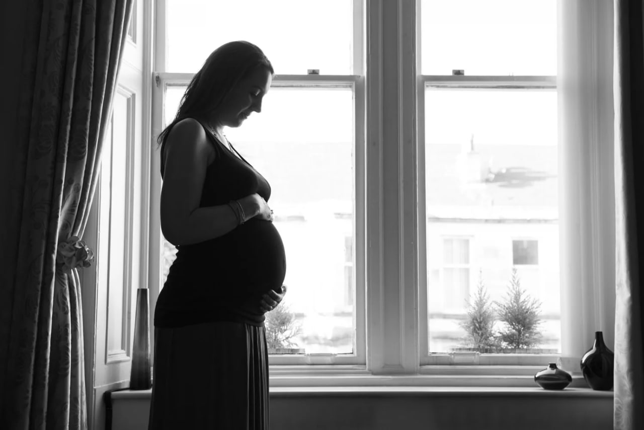 A pregnant woman stands in silhouette by a bright window during a maternity session at Maxwell Park in South Lanarkshire