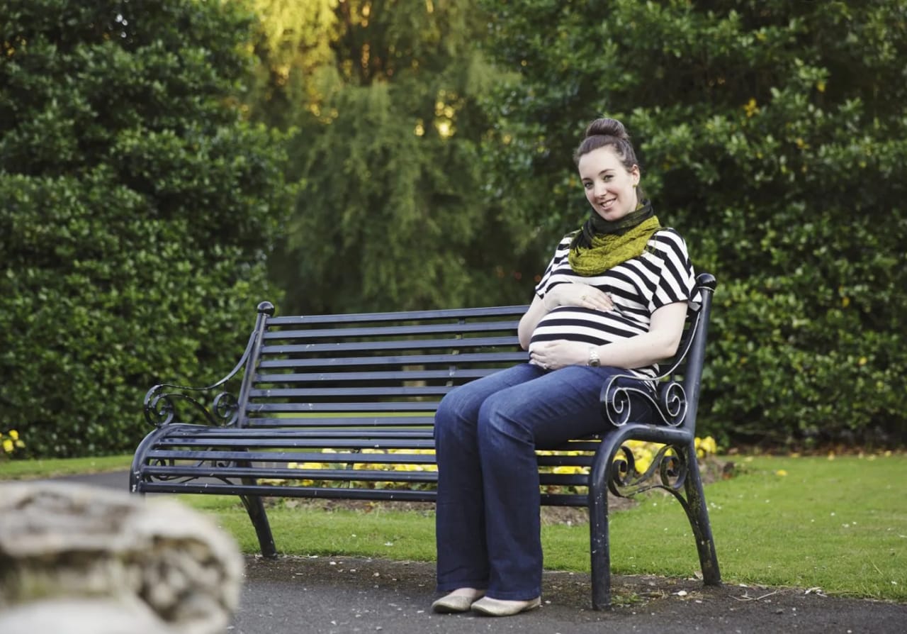 A pregnant woman relaxes on a bench during a maternity session at Maxwell Park, surrounded by greenery.