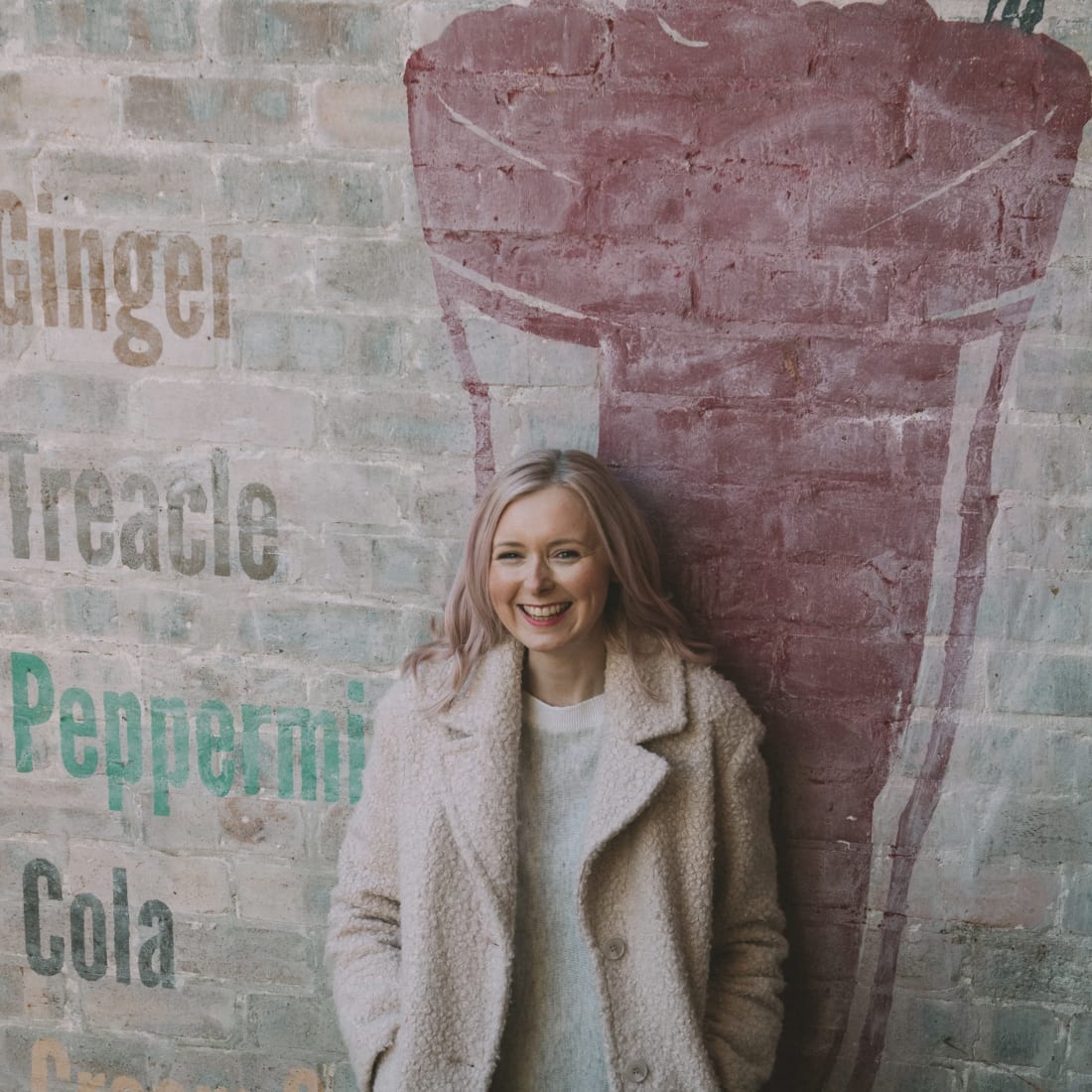 Photographer Louise Mallan standing in front of a vintage drink mural on a brick wall, wearing a coat.