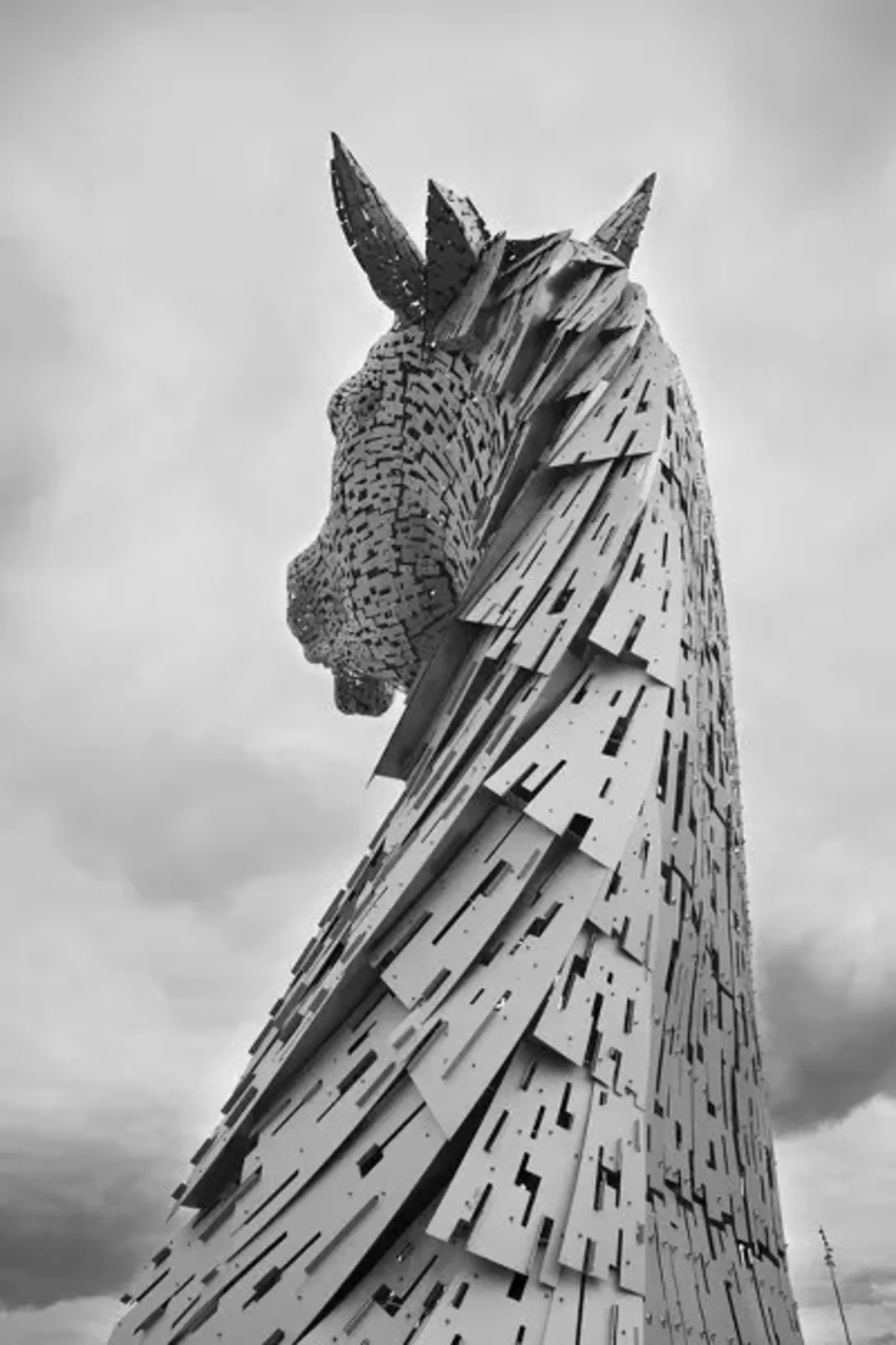 A side view of the towering Kelpies monument against a cloudy sky, highlighting its intricate metal design
