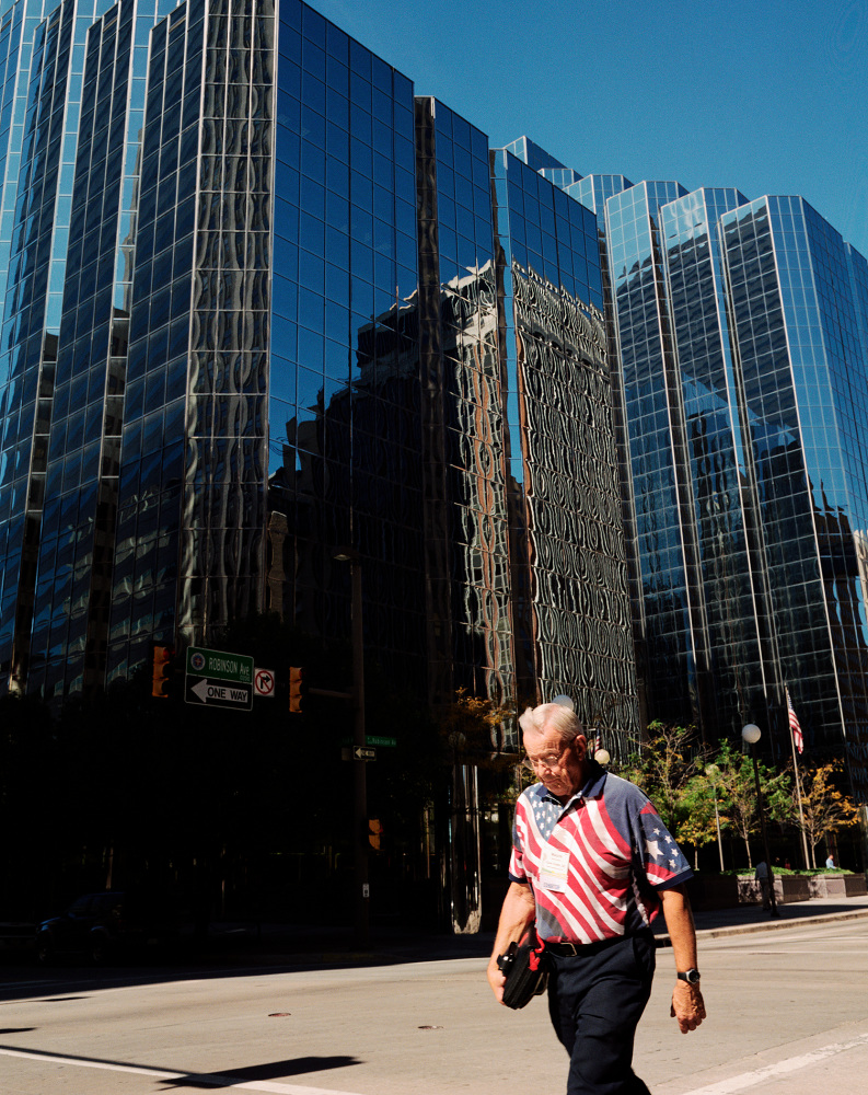 A man wearing a patriotic shirt walks past modern glass skyscrapers
