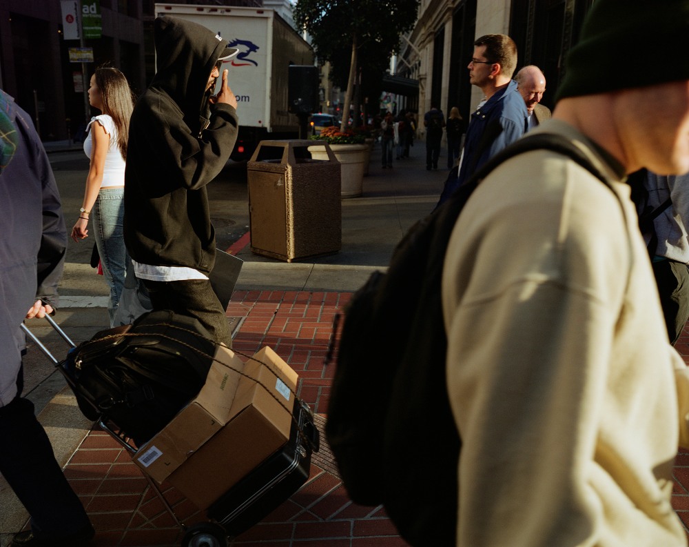 A person in a black hoodie pulls a cart with boxes while others walk by on a busy street
