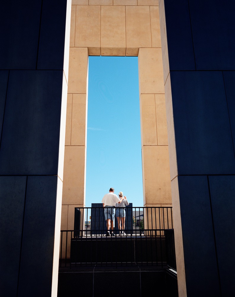Two people standing on a balcony framed by a large open structure against a clear blue sky