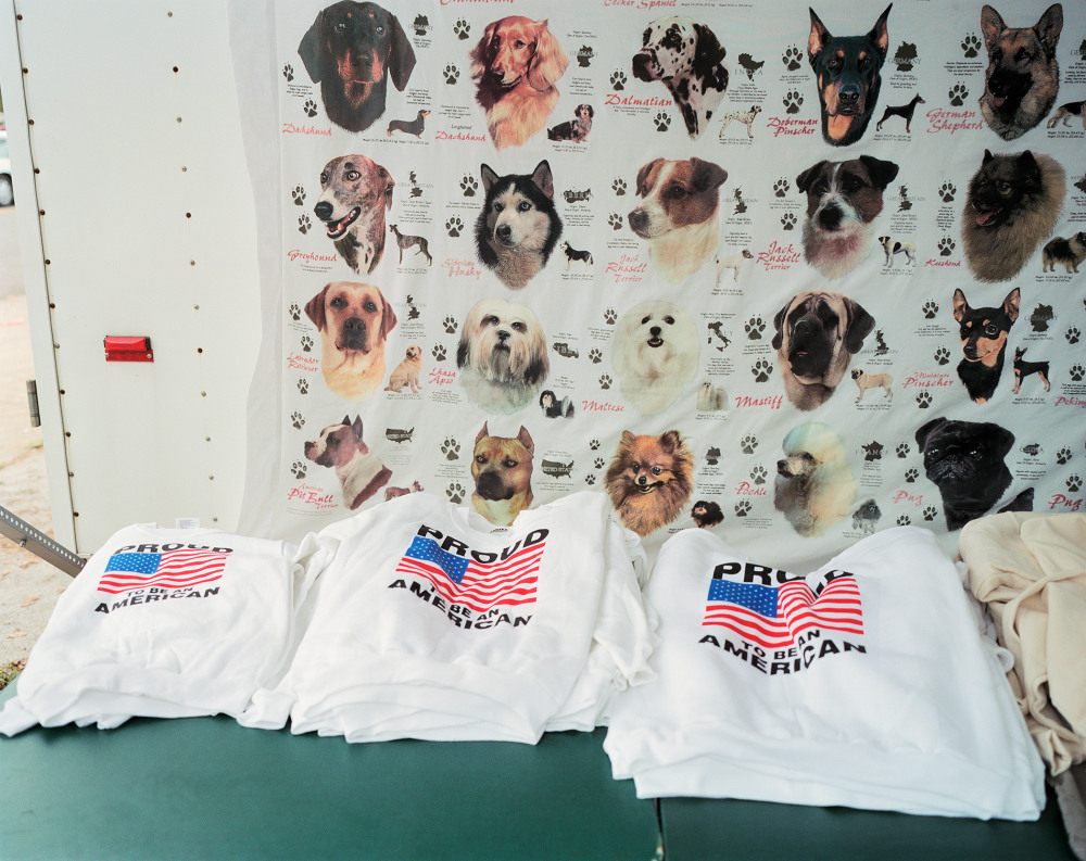 Three white sweatshirts with "PROUD AMERICAN" text and American flags displayed on a table in front of a backdrop of various dog images