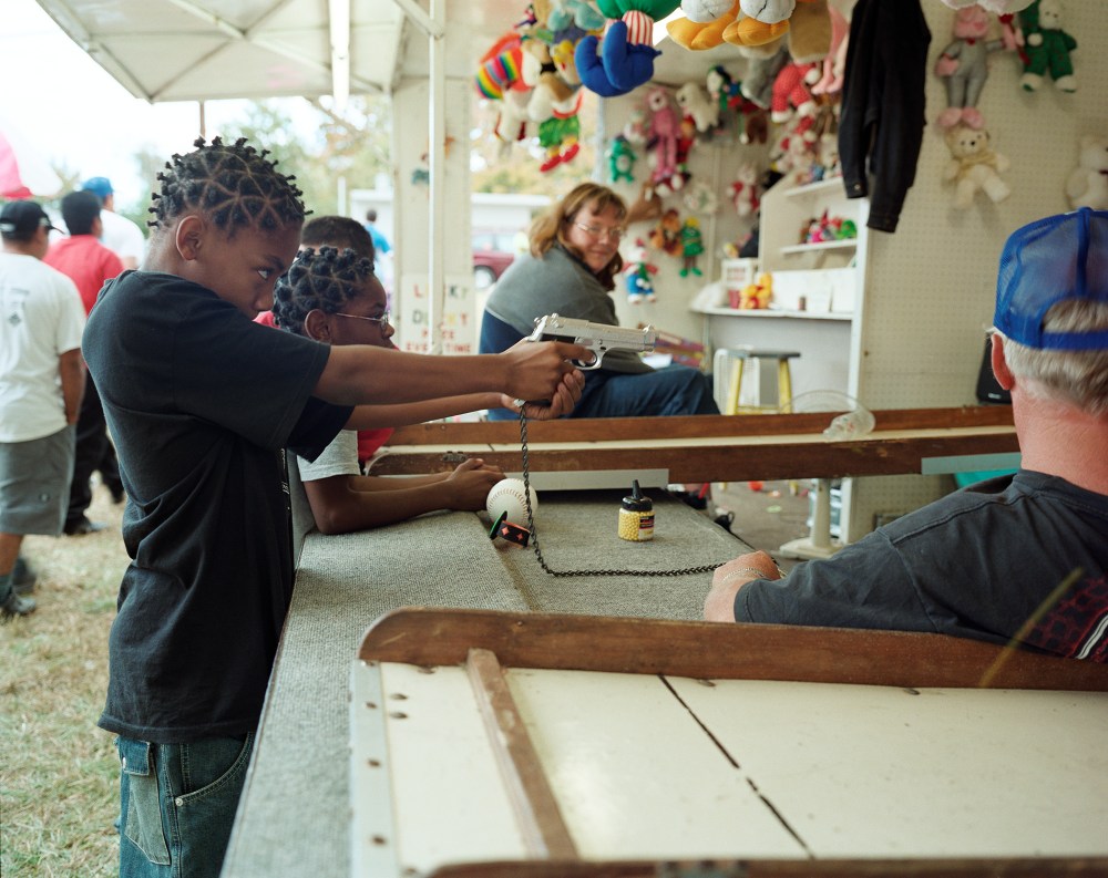 A child aims a toy gun at a game booth while other children and adults observe