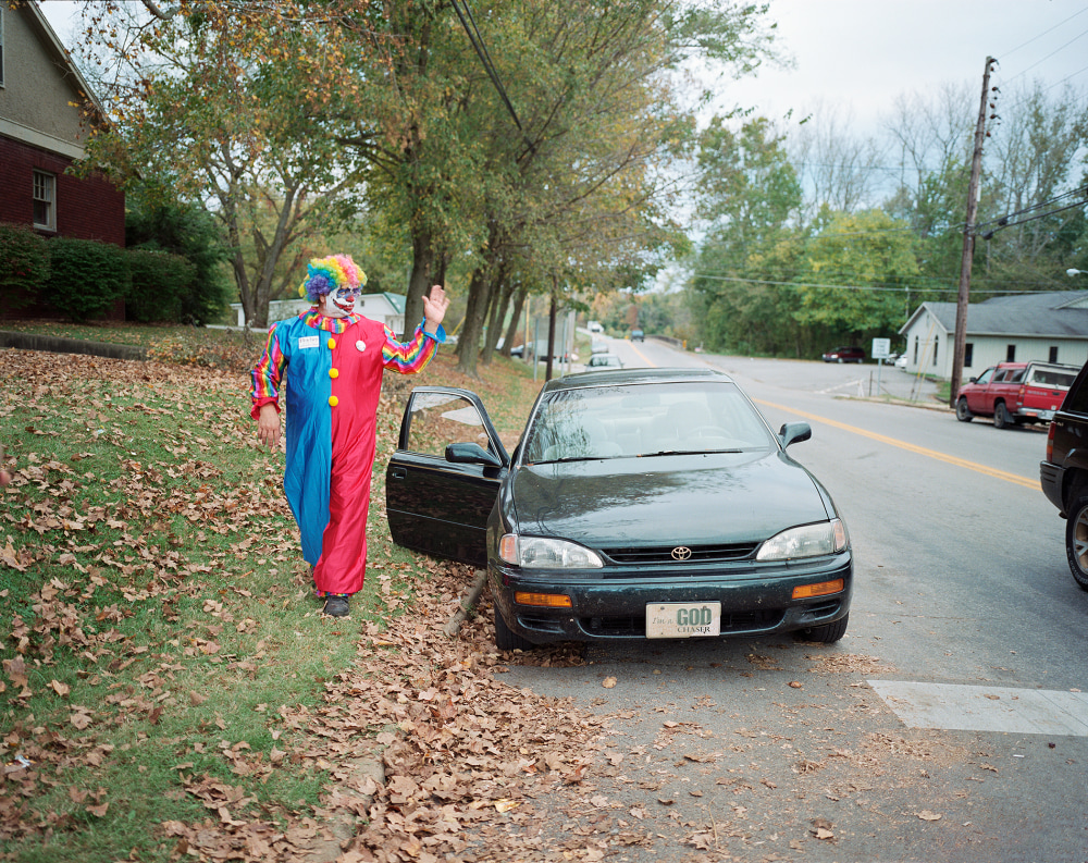 A clown in colorful attire waves beside a black car on a leaf-covered roadside