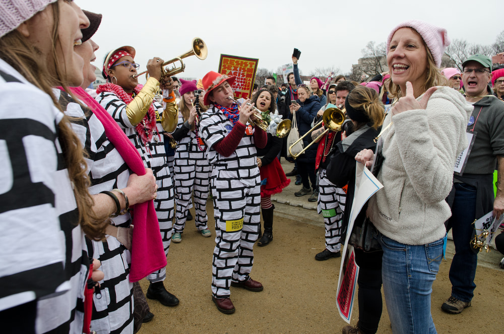 A lively crowd of musicians in striped outfits playing instruments while engaging with cheerful protesters