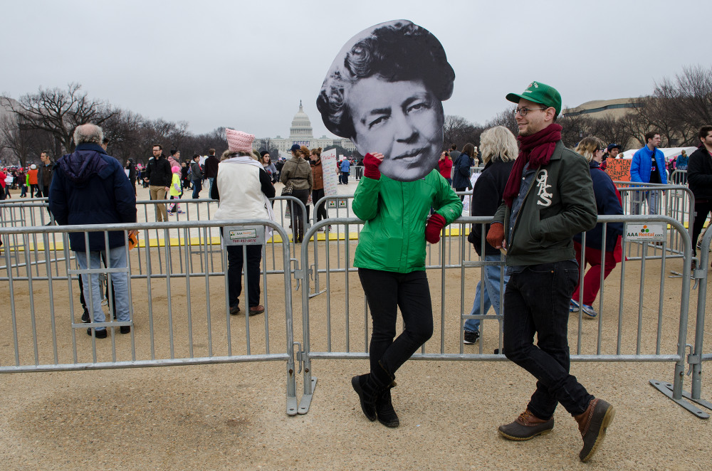 A person in a green shirt holds a large cutout of Eleanor Roosevelt at a crowded outdoor event near the Capitol