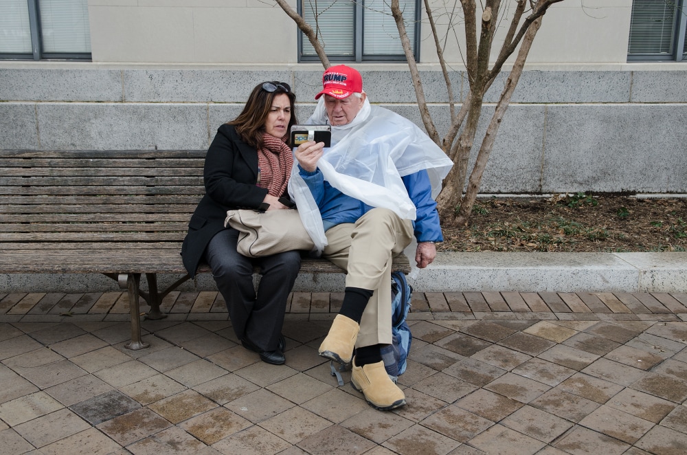 A man in a red cap and a plastic poncho sits on a bench with a woman, both looking at a phone