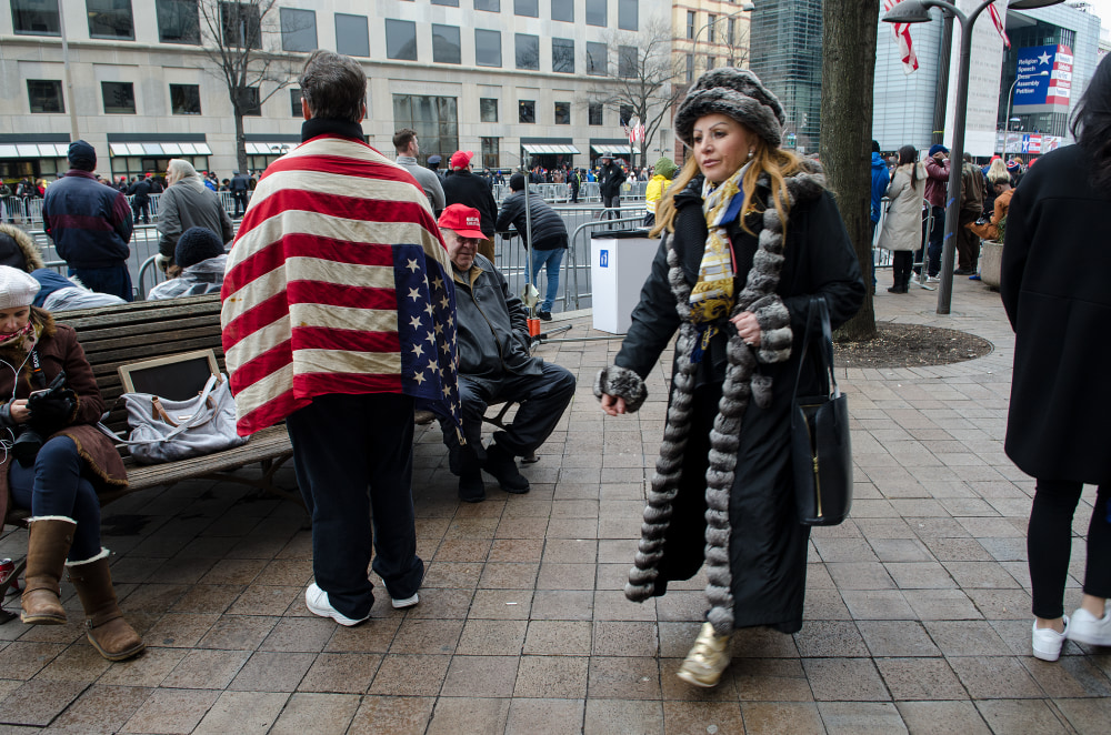 A woman in a fur coat walks past people in winter attire, including one wrapped in an American flag