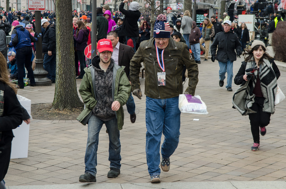 Two men in casual clothing walk through a crowded outdoor area with people in the background