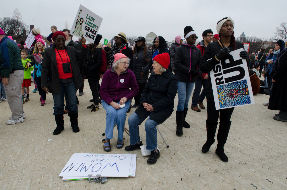 A diverse group of protesters, including two seated elderly women, holding signs at a rally