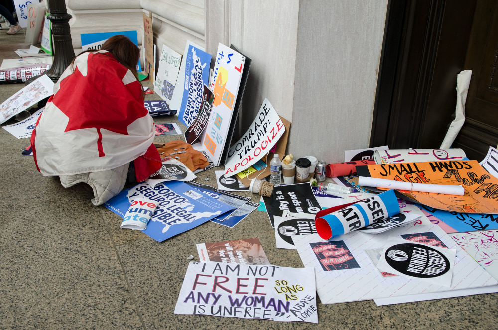A person in a red and white outfit sits on the ground surrounded by protest signs