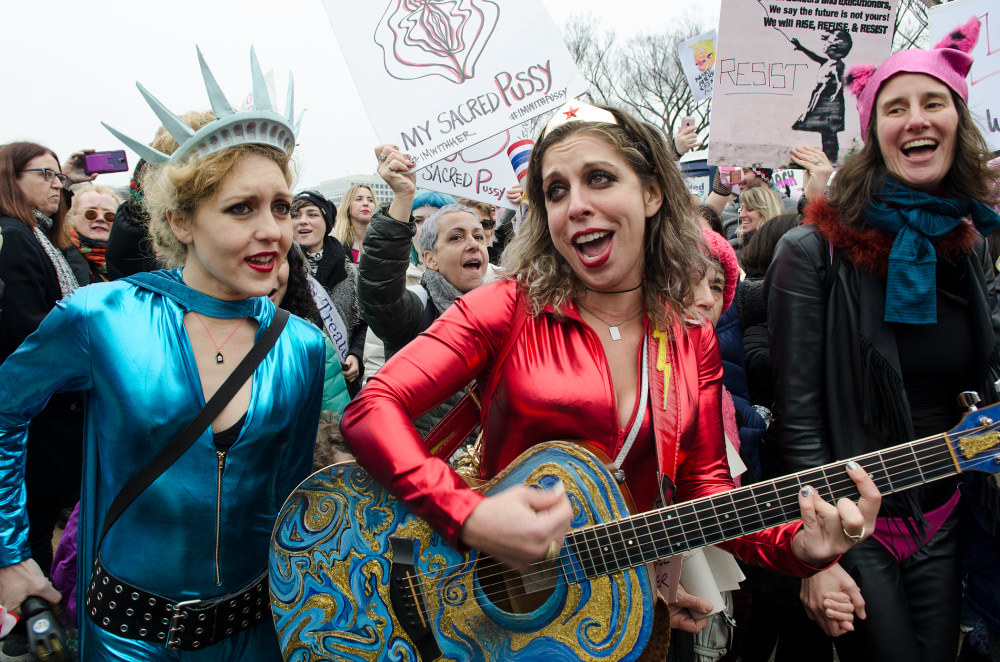 Two women in colorful costumes sing and play guitar at a lively protest with supporters in the background
