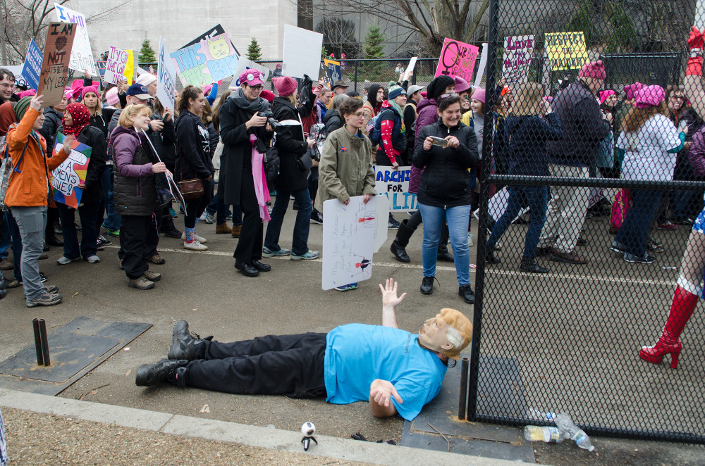 A crowd of protesters with signs, some wearing pink hats, with a mannequin resembling a person lying on the ground in front of a fence