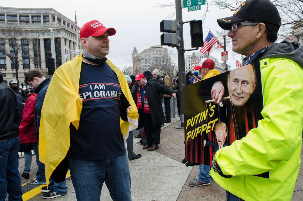 Two men engage in a political protest, one wearing a "Deplorable" shirt and the other holding a sign with Putin's image