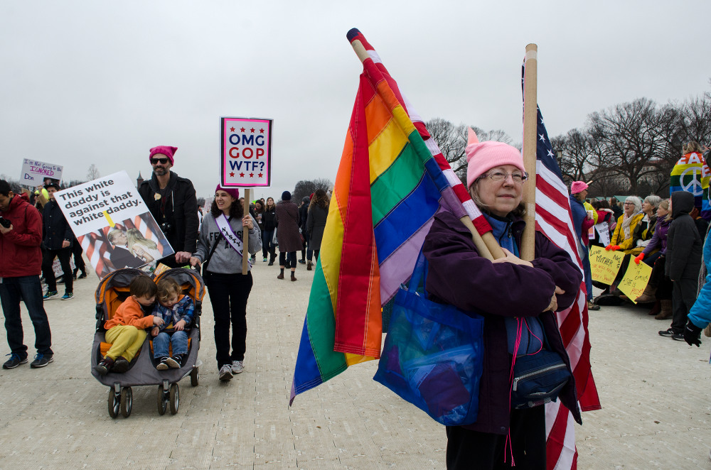 A diverse group of protesters marching, holding signs and flags, including a rainbow flag and an American flag
