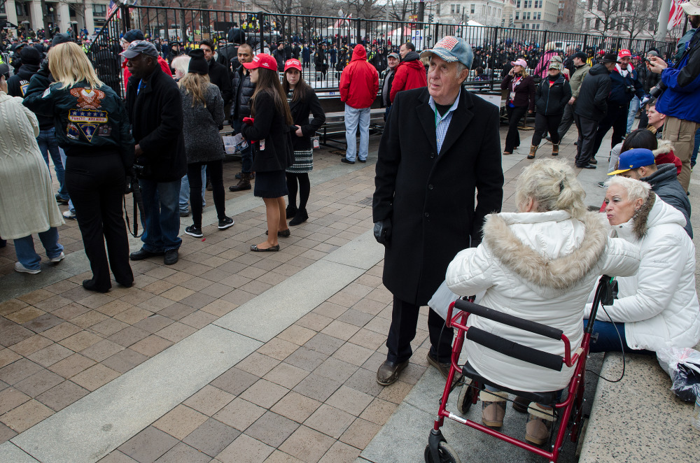 A crowd of people, some wearing red hats, gathers in an outdoor setting with a man in a coat standing nearby