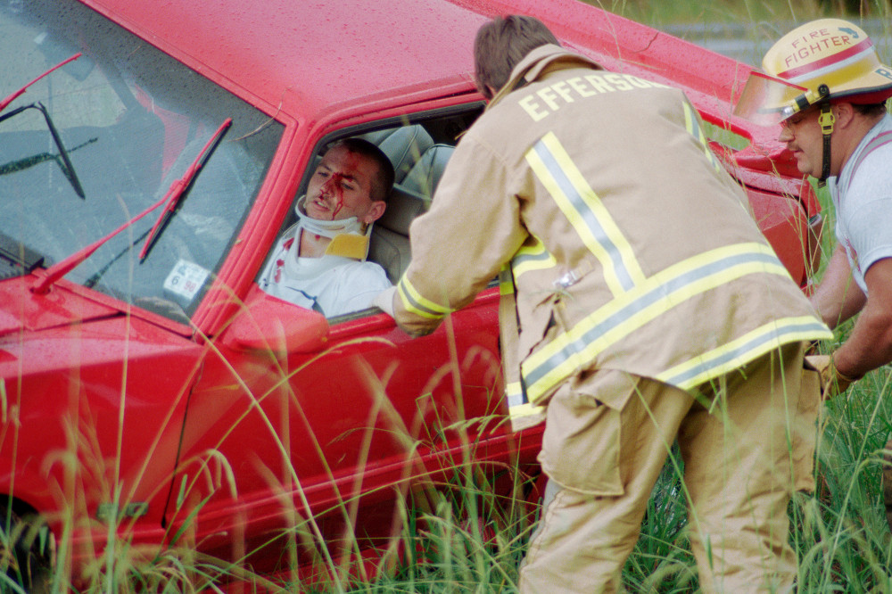 Firefighters assisting an injured person trapped in a red car in a grassy area